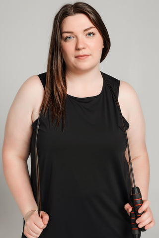 Well Fit Activewear. Female model wears long length, drop-arm gym vest and black compression leggings to the ankle. Well Fit tops and leggings are available in UK sizes 8 to 30. Free shipping over £100. Grace stands face on to camera with a black handle skipping rope placed around her shoulders.