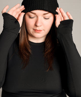 Well Fit Activewear. Female model wears black compression gym top in a longer length tunic style and black compression leggings to the ankle. Well Fit tops and leggings are available in UK sizes 8 to 30. Free shipping over £100. Grace stands straight on to camera, looking down to the floor. She is wearing a black, woollen beanie hat which she is adjusting with both hands therefore revealing the thumbhole design of the cuff top.