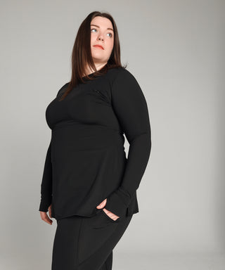 Well Fit Activewear. Female model wears black compression gym top in a longer length tunic style and black compression leggings to the ankle. Well Fit tops and leggings are available in UK sizes 8 to 30. Free shipping over £100. Grace stands side on to camera with hand in her leggings pocket to reveal the top cuff detail.